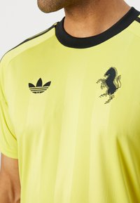 Žlté športové tričko s čiernymi akcentmi. Obsahuje logo Adidas a emblém koňa. Hladký textil, okrúhly výstrih a pruhovaný dizajn.