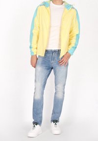 Windbreaker-jas in geel met blauwe accenten, voorzien van een rits, trekkoordkap en elastische boorden. Draagt een lichte blauwe spijkerbroek en witte sneakers.