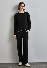 Svart stickad tröja med rundad hals och vita ribbade manschetter, kombinerad med svarta byxor och vita sneakers på en strukturerad yta.