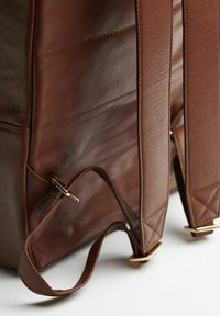 Ruskea nahkainen reppu, jossa on tikatut olkahihnat ja kultaiset metalliosat. Karkea pinta, jossa näkyvät saumurikeskitykset.