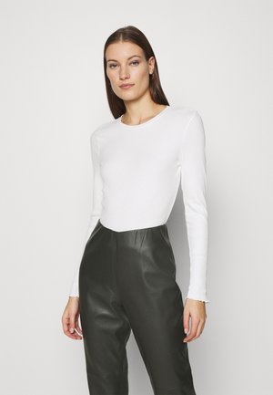 Bílý dlouhý rukáv ribbovaný top v kombinaci s tmavě zelenými koženými kalhotami s vysokým pasem, které mají hladkou texturu a přiléhavý design.