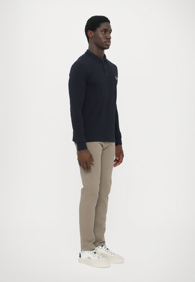 Polo de manga longa azul-marinho com colarinho, calças bege e ténis brancos. Apresenta um pequeno logótipo no peito e um design ajustado.