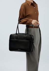 Schwarze Lederhandtasche mit goldfarbenen Beschlägen, doppelten Griffen und einer strukturierten Form. Auffällige Details sind die vordere Reißverschlusstasche und das Logo.