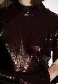 Bordeaux geklede top met een hoge hals, met een gladde textuur en een glanzend, reflecterend oppervlak. Close-upweergave benadrukt het intrigerende pailettenpatroon.