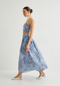 Helles blaues Zweiteiler-Outfit mit einem taillierten Oberteil und einem langen, fließenden Rock mit floralem Muster und plissierter Gestaltung. Die Materialien wirken leicht.