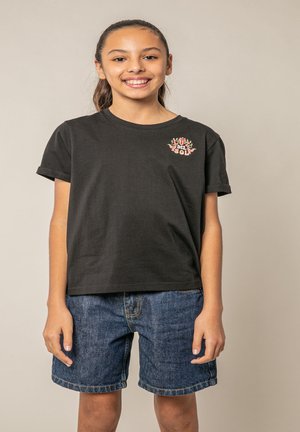 Fille souriante avec une queue de cheval portant un t-shirt noir "Luz del Sol" et un short en jean, debout devant un fond beige uni.