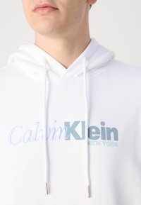 Biała bluza z kapturem z logo z przodu, na którym widnieje napis "Calvin Klein" w jasnoniebieskim i ciemnoniebieskim kolorze, z zakrzywionymi sznurkami do ściągania oraz srebrnymi akcentami.