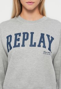 Šedý sveter so širokým námořníckym modrým nápisom "REPLAY" na hrudi, okrúhlym výstrihom a elastickými manžetami. Jemná textúra tkaniny je viditeľná.