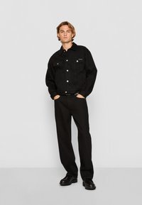 Black denim jacket with silver buttons, two chest pockets, and black trousers. Smooth fabric with a relaxed fit and minimalistic design.