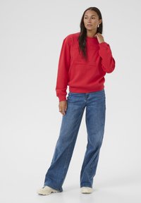Czerwona bluza z nadrukiem z przodu, luźny krój, ribbowe mankiety, w połączeniu z szerokimi niebieskimi dżinsami i kremowymi sneakersami. Gładka tekstura, casualowy styl.
