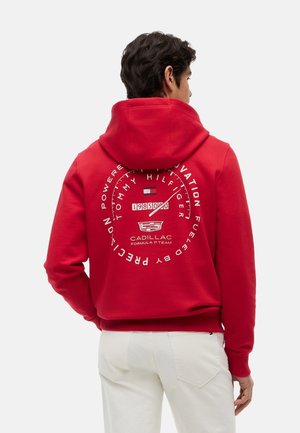 Uomo che indossa una felpa con cappuccio rossa con una grafica circolare bianca di un contagiri e il testo "Tommy Hilfiger", "Cadillac Formula 1 Team" sulla schiena.