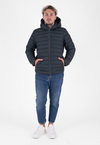 Giovane con le mani in tasca, indossa un piumino nero con cappuccio, jeans blu e sneakers bianche, in piedi su uno sfondo bianco.