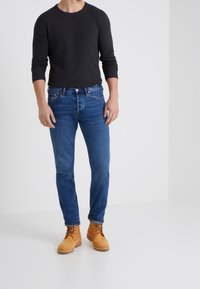 Mörk långärmad tröja, blå jeans och beige arbetsskor. Jeans har klassisk femficksdesign. Tyget verkar slätt med lätt textur.