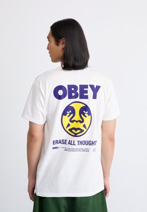 Hombre con cabello largo que lleva una camiseta blanca con el texto púrpura "OBEY", un gráfico de cara amarilla y púrpura y el lema "ERASE ALL THOUGHT" en la parte de atrás.
