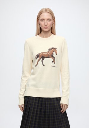 Femme aux cheveux blonds raides portant un pull crème avec un motif cheval marron et une jupe à carreaux foncée, debout devant un fond uni.