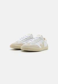 White canvas sneakers with beige suede accents, textured rubber sole, and stitched detailing. Features flat laces and a logo on the tongue.