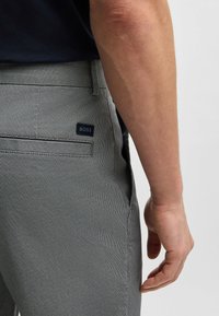 Gray textured pants with a subtle pattern, featuring a BOSS logo tag on the back pocket, shown beside a hand.