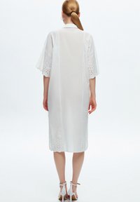 Robe blanche mi-longue avec des manches courtes évasées. Présente une broderie à jour au niveau de l'ourlet et un design droit. Tissu léger.