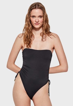 Bañador negro sin tirantes con fruncidos y lazos laterales. Textura suave y piernas de corte alto, con acentos dorados en los extremos de los lazos. Diseño simple.