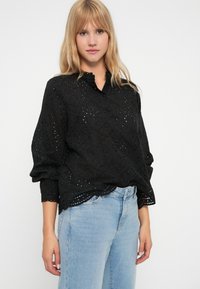 Svart bomullsbloyse med perforeringsdetaljer, avslappet passform og bølget kant. Modellen er kombinert med lyseblå denimjeans.