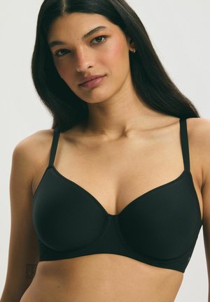 Young woman with long dark hair wearing a fitted black bra, facing forward with neutral expression against plain background.