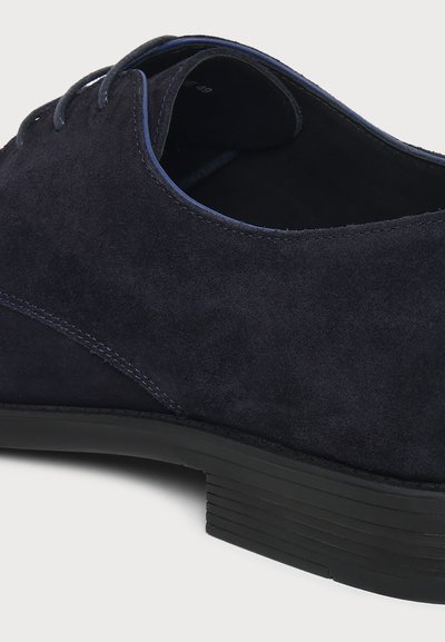 Gros plan d'une chaussure habillée en daim bleu marine foncé avec fermeture à lacets et talon empilé noir sur fond clair.
