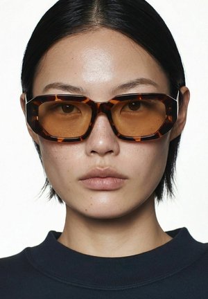 Personne aux cheveux noirs raides portant des lunettes de soleil oversize à monture écaille de tortue avec des verres ambrés et un haut sombre sur fond blanc uni.