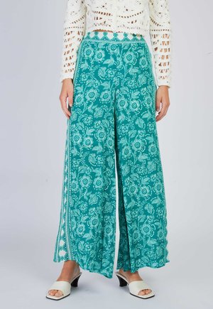 Kobieta nosząca szerokie spodnie w kolorze zielonkawo-niebieskim z białym kwiatowym wzorem, zestawione z białymi sandałami na obcasie oraz białą szydełkowaną bluzką z długim rękawem.