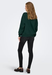 Sweatshirt verde oversize con scollo a girocollo e polsini a costine, abbinato a jeans skinny neri con dettagli strappati e scarpe scure.
