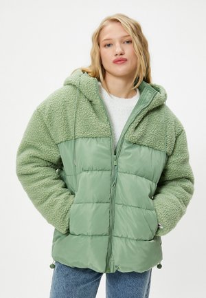 Junge Frau trägt eine grüne, mit Kapuze versehene Steppjacke mit strukturierten Ärmeln und den Händen in den Taschen, steht vor einem schlichten weißen Hintergrund.