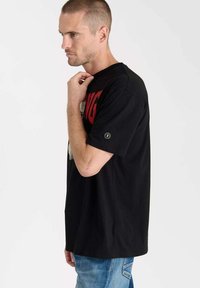 Camiseta de algodón negra con un estampado gráfico rojo. Corte relajado, mangas cortas y un pequeño detalle de logotipo en la manga izquierda.