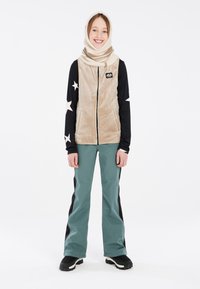 Flauschige beige Weste mit Reißverschluss, schwarzes Langarmshirt mit weißen Sternen, grüne Skihose und schwarze Stiefel. Ein warmer Hals-Schal ergänzt das Outfit.