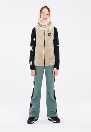 Fuzzy beige vest met rits, zwart shirt met lange mouwen met witte sterren, groene skibroek en zwarte laarzen. Warme nek sjaal completeert de outfit.