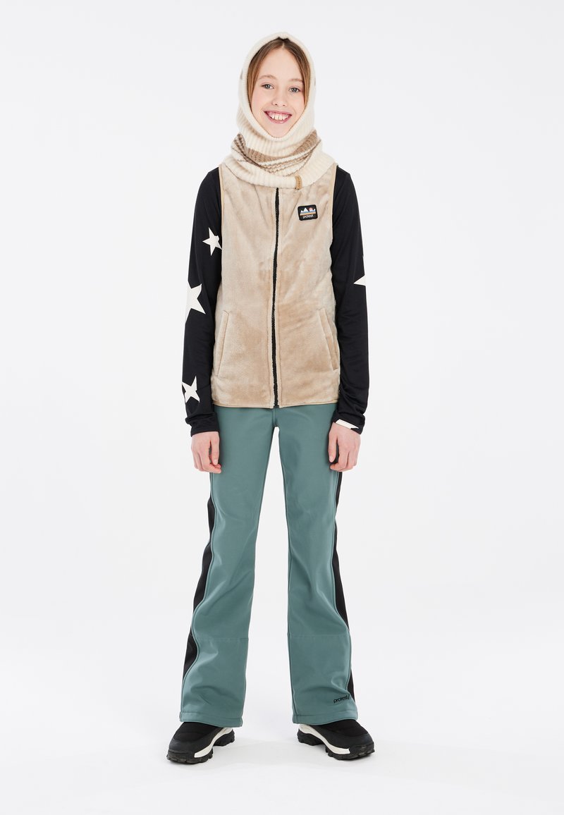 Flauschige beige Weste mit Reißverschluss, schwarzes Langarmshirt mit weißen Sternen, grüne Skihose und schwarze Stiefel. Ein warmer Hals-Schal ergänzt das Outfit.