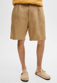 Pantalones cortos de algodón beige con corte recto, frente con plegados y bolsillos laterales, combinados con zapatos beige tipo slip-on. Textura suave, diseño casual.