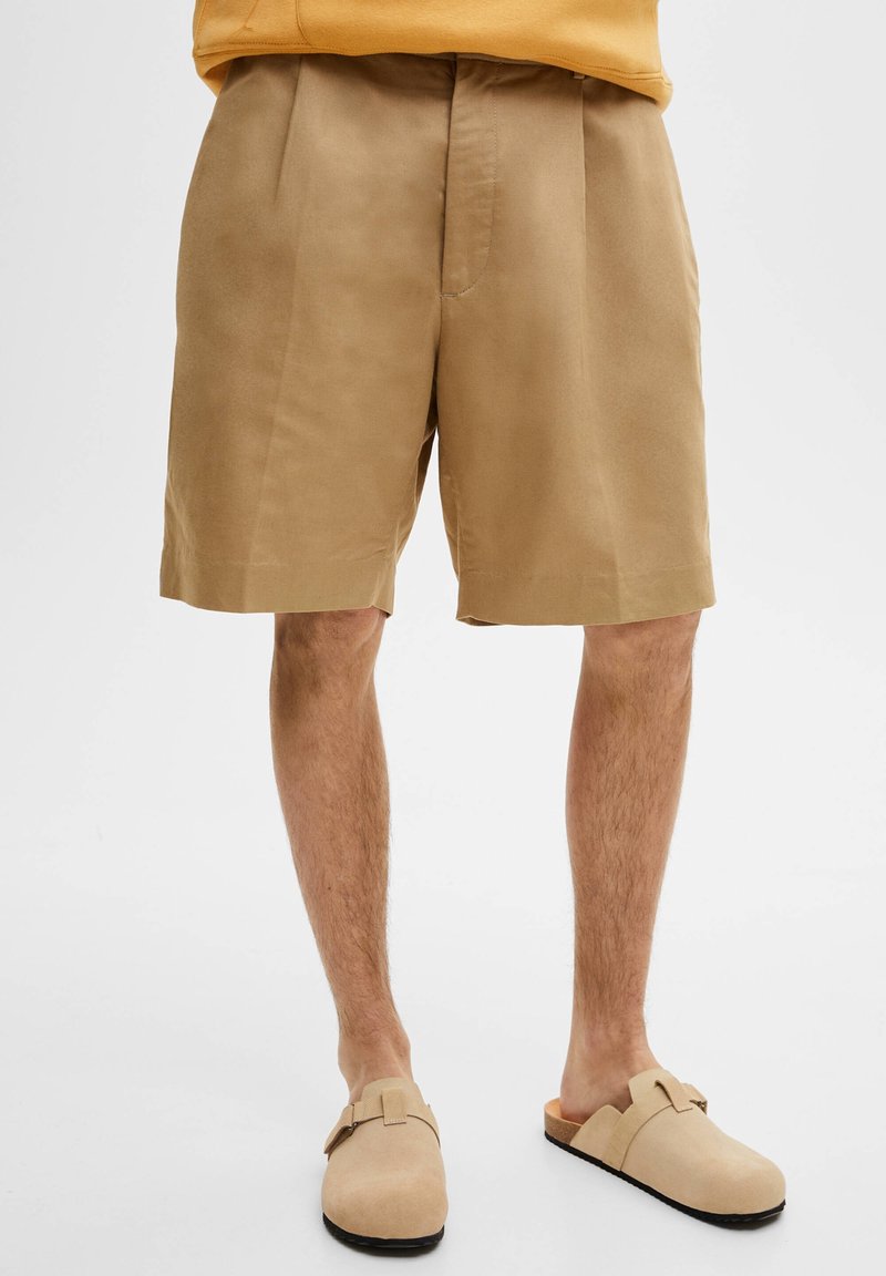Pantalones cortos de algodón beige con corte recto, frente con plegados y bolsillos laterales, combinados con zapatos beige tipo slip-on. Textura suave, diseño casual.