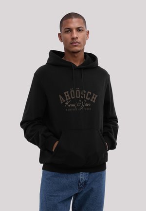 Black hoodie with a front pocket, featuring a brown graphic text design. The material is soft with ribbed cuffs and hem. Worn with blue jeans.