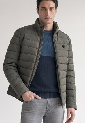 Mann trägt eine grüne gesteppte Jacke über einem blauen Strickpullover mit grauer Jeans, steht mit einer Hand in der Tasche vor einem schlichten Hintergrund.