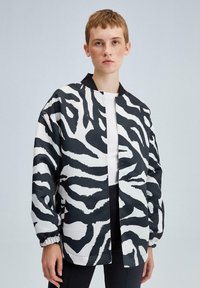 Črna in bela jakna z izrazitim zebra vzorcem, sproščenega kroja, z elastičnimi manšetami in dvignjenim ovratnikom. Nošena preko belega vrha.
