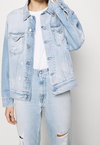 Giacca di jeans blu chiaro con tasche frontali, cuciture visibili, chiusura con bottoni e dettagli usurati, abbinata a jeans a vita alta coordinati.