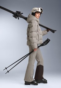 Bubasto jakna s uzorkom ptičjeg zuba, u kombinaciji s odgovarajućim širokim hlačama, drži skije i skijaške štapove, s bijelim kacigom i naočalama.