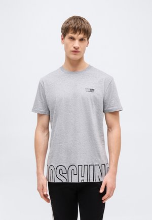 Jonge man met kort bruin haar, gekleed in een lichtgrijs Moschino T-shirt met grote zwarte logotekst aan de zoom en een klein logo op de borst.