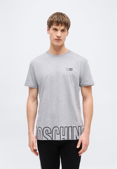Jeune homme aux cheveux bruns courts portant un t-shirt gris clair Moschino avec un grand lettrage noir du logo à l'ourlet et un petit logo sur la poitrine.