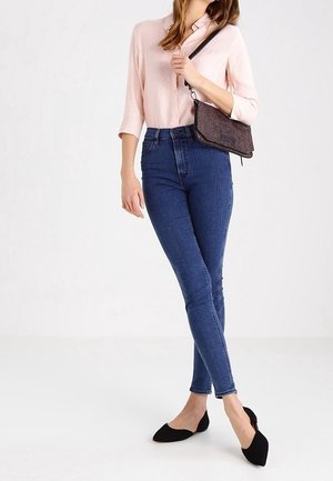 Lichtroze overhemd met opgerolde mouwen, donkerblauwe skinny jeans, zwarte puntige platte schoenen en een kleine, gestructureerde crossbodytas.