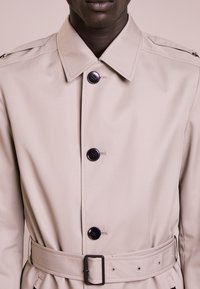 Trench coat beige con collo a camicia, bottoni neri frontalmente, cintura con nodo automatico e spalline decorative sulle spalle.