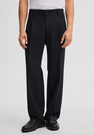 Mann trägt schwarze plissierte Hose mit Knopf- und Reißverschluss, kombiniert mit schwarzen Leder-Loafers, steht vor einem schlichten Hintergrund.