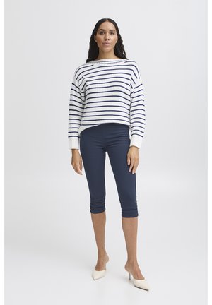 Pikajuuste ja tumedate juustega naine, kes kannab valget ja tumesinist triibulist kampsunit, tumesiniseid lühikesi pükse ja valgeid kontsakingi, seisab ühevärvilise tausta ees.