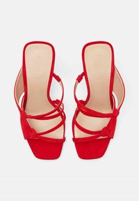 Sandales à talons hauts en daim rouge avec un bout ouvert, présentant des lanières croisées et de petits nœuds accents au niveau de la cheville. Design à enfiler carré.