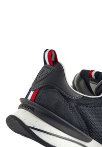 Zapatilla negra que presenta materiales de ante y malla, con una entresuela blanca con acentos salpicados y una lengüeta en el talón a rayas en rojo, blanco y azul.