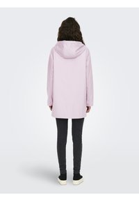 Chaqueta impermeable rosa claro con capucha, mangas largas y corte recto, combinada con pantalones oscuros de corte slim y calzado negro.